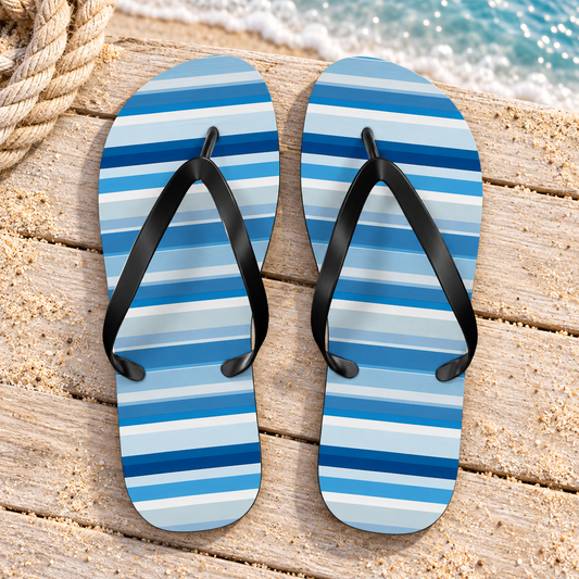Shades of Light Blue Stripes Flip Flops, product-only overhead view with the printed pattern clearly visible. Setting: pale sandy beach boardwalk with sea-blue water edge.