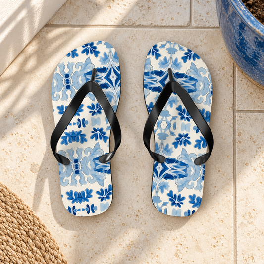 Blue Spanish Tile Flip Flops, product-only overhead view with the printed pattern clearly visible. Setting: cream coastal balcony tiles beside a blue ceramic pot edge.