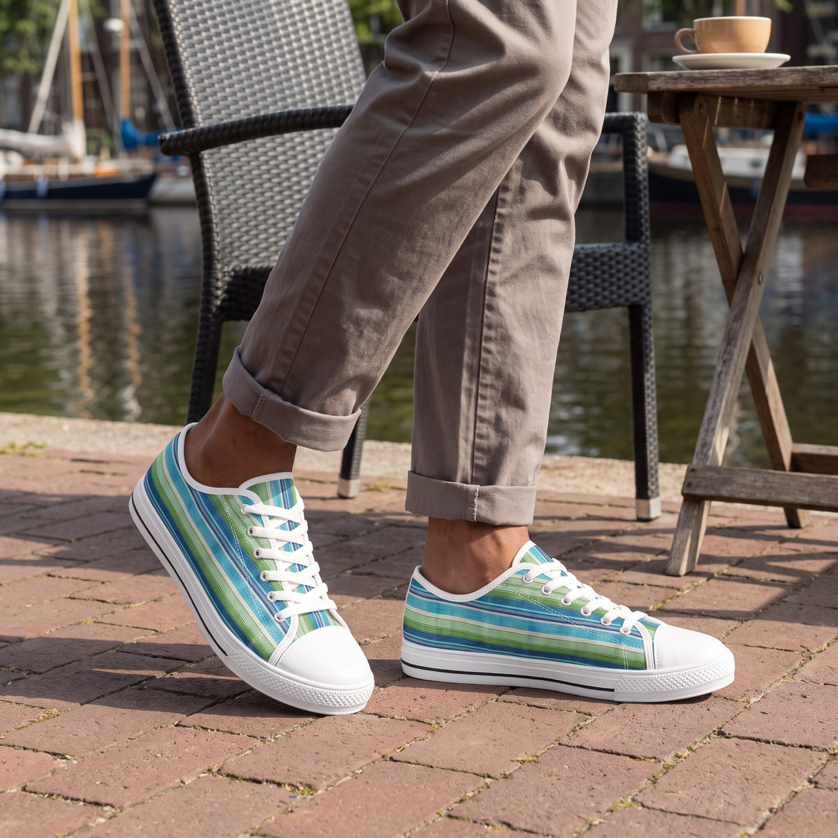 Ocean Whisper Stripes Men's Low Top Sneakers. Lifestyle scene: Canal towpath café terrace; dry brick, charcoal woven chair, soft sailboat reflections
