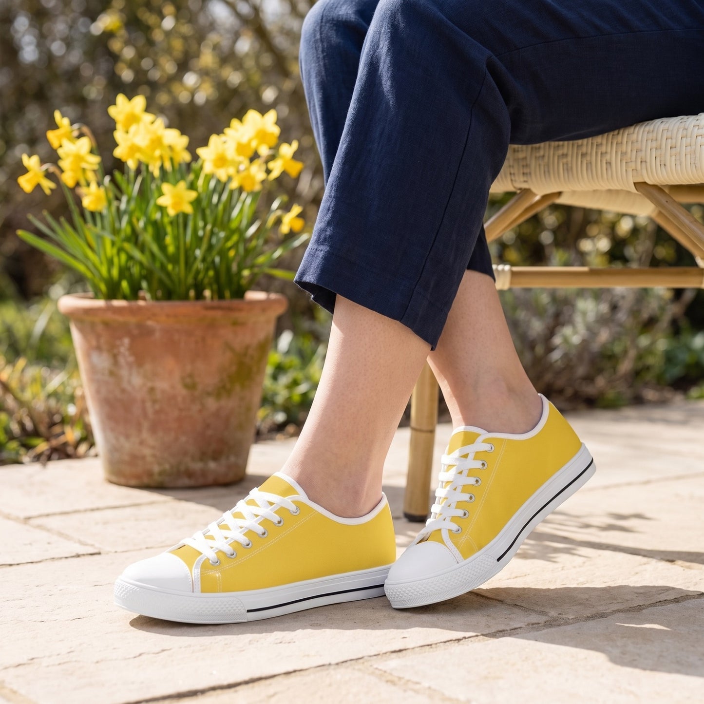 Daffodil Ladies' Two-Tone Low Top Sneakers. Lifestyle scene: Garden breakfast terrace; pale limestone, daffodil pot behind chair, cream woven seat