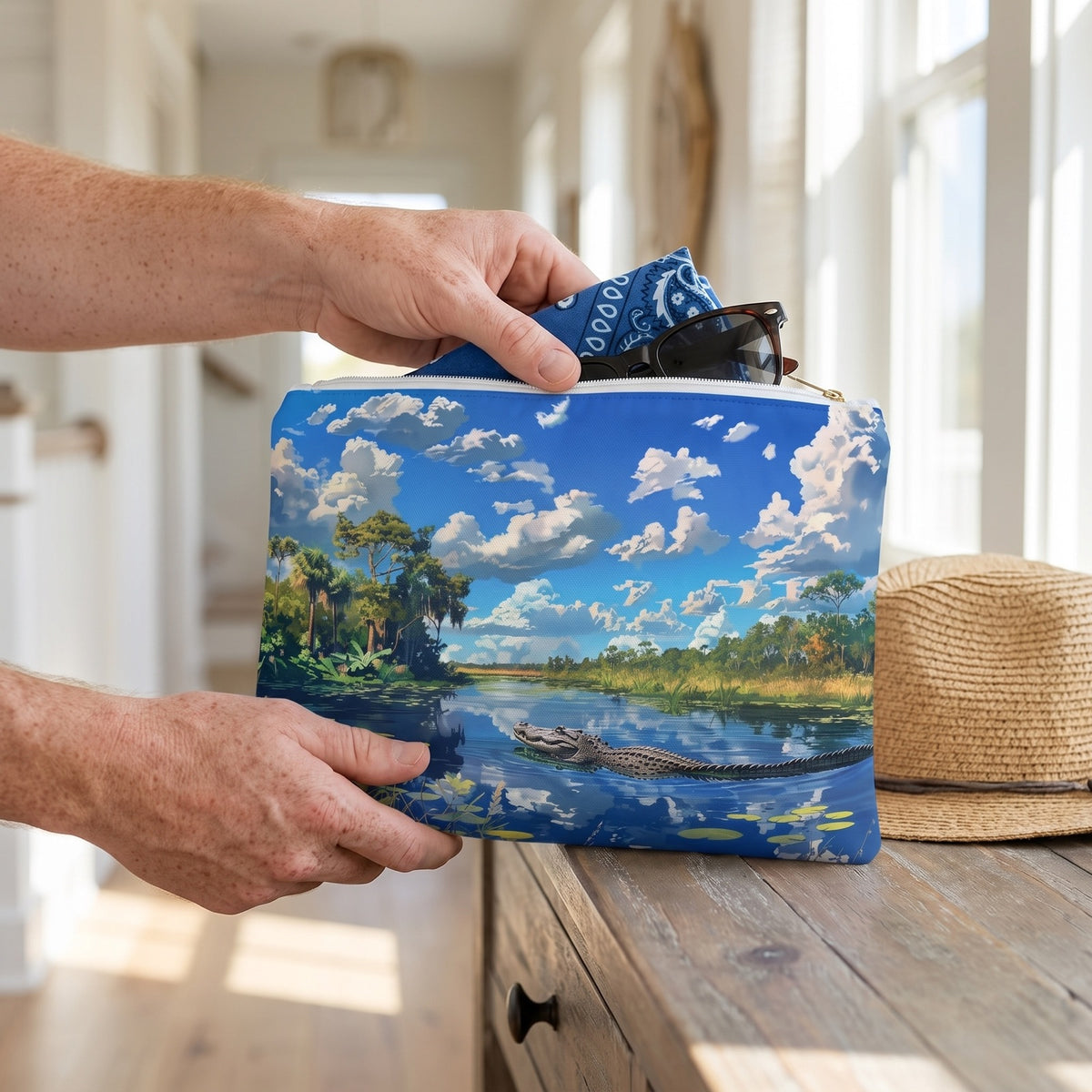 Large Everglades Encounter accessory pouch (11.81 × 8.35 inches) in use: man packs a folded bandana and sunglasses into pouch beside a straw hat. Lifestyle mockup with hands showing scale.