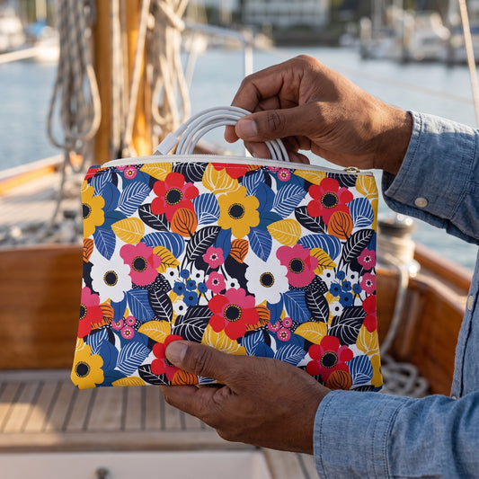 Small Blossom Burst accessory pouch (8.35 × 5.98 inches) in use: man in a chambray shirt slips a coiled charging cable into the pouch held above his knee. Lifestyle mockup with hands showing scale.