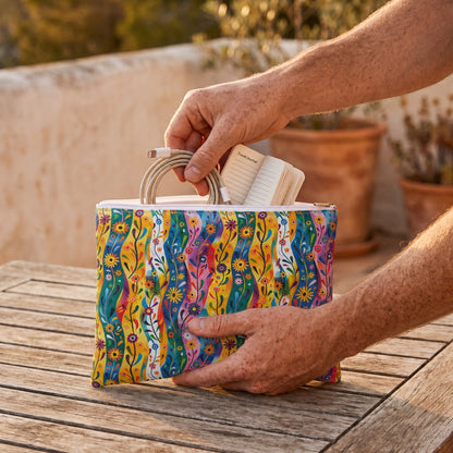 Large Floral Rhythms accessory pouch (11.81 × 8.35 inches) in use: man packs charging cable and a small paperback-size travel journal into pouch. Lifestyle mockup with hands showing scale.