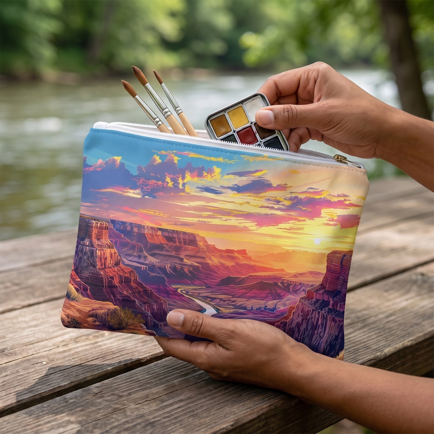 Large Golden Grand Canyon Glow accessory pouch (11.81 × 8.35 inches) in use: adult watercolor hobbyist places a small tin and three brushes into pouch, river blurred beyond. Lifestyle mockup with hands showing scale.