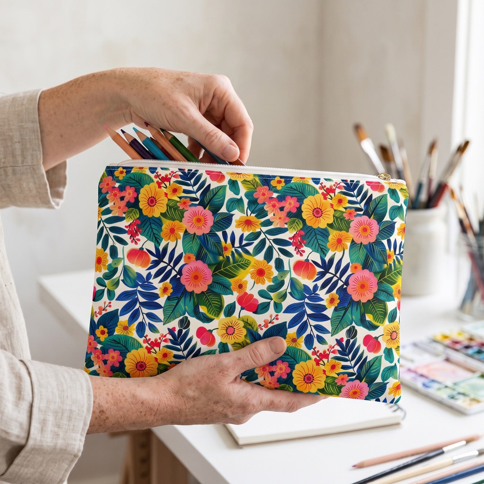 Large Blossom Bliss accessory pouch (11.81 × 8.35 inches) in use: woman in oatmeal linen gathers six colored pencils into the pouch, sketchbook just behind. Lifestyle mockup with hands showing scale.