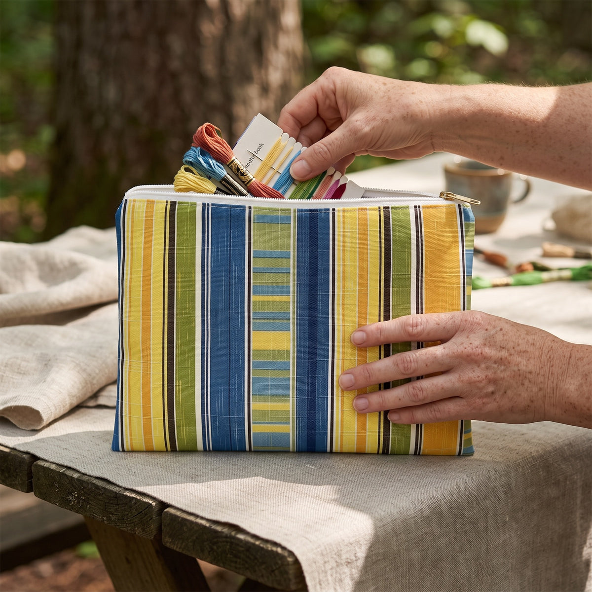 Large Summer Stripes Delight accessory pouch (11.81 × 8.35 inches) in use: woman packs embroidery floss and a small needle book into pouch. Lifestyle mockup with hands showing scale.