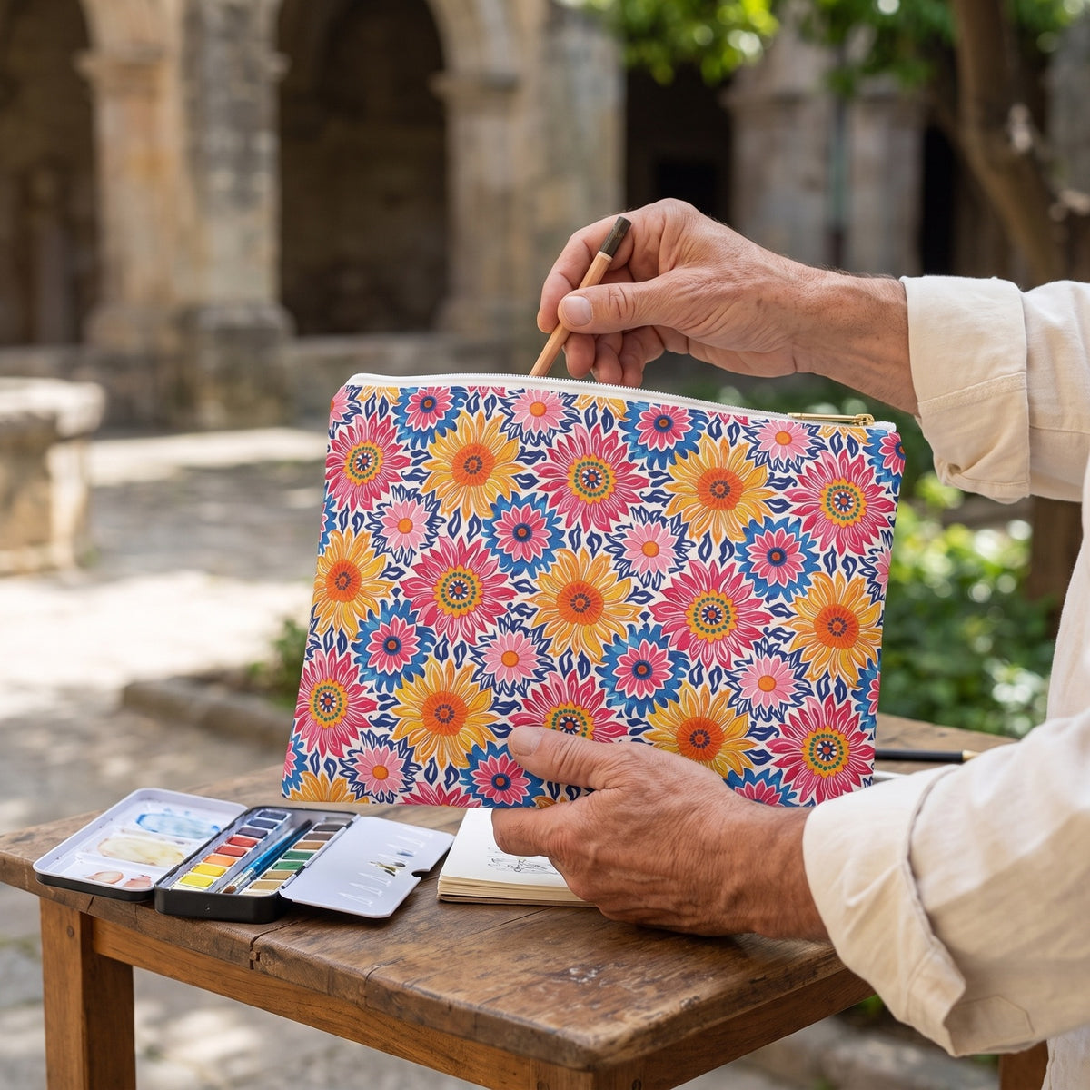 Large Sunsplash Petals accessory pouch (11.81 × 8.35 inches) in use: man with rolled cream sleeves selects a pencil from the open pouch beside a tiny watercolor set. Lifestyle mockup with hands showing scale.