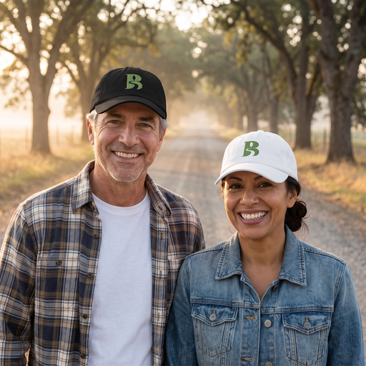 A smiling man in flannel wears the black Bliss Road Studios embroidered logo cap and a smiling woman in a denim jacket wears the white one, walking a misty oak-lined country road at sunrise
